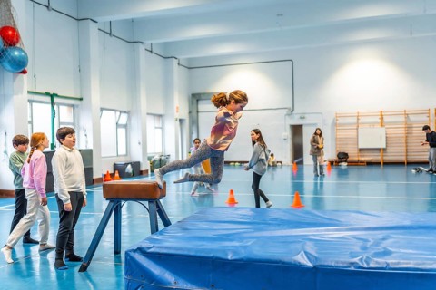 Elementary school girl practicing gymnastics, jumping over a pommel horse in the school gym watched