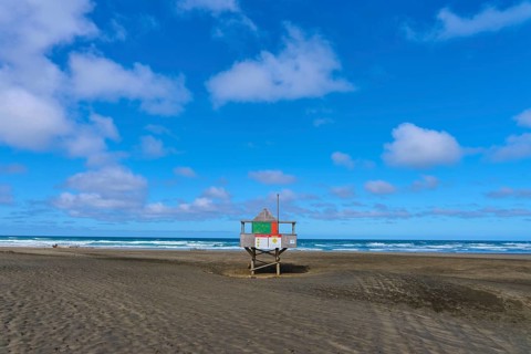 A rescue tower stands alone on a wide sandy beach under a blue sky, Bethells Beach, Te Henga,