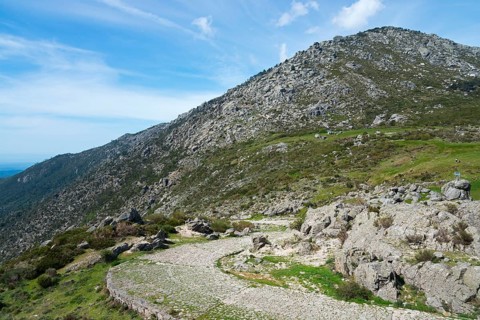 A winding road leads through a rocky landscape with green vegetation and high mountains in the