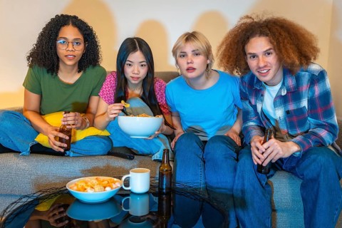 Four young friends enjoying a night in, watching television, eating snacks and drinking beer,