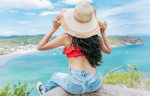 Rear view tourist girl sitting at a viewpoint looking at the beach. Back view of woman on vacation