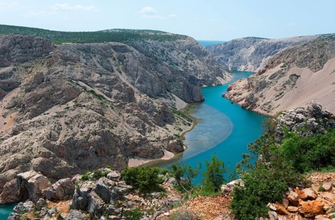 A deep gorge with a turquoise-blue river, surrounded by barren rocks, under a summer sky,