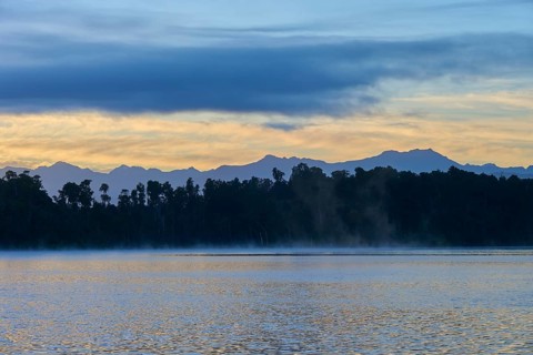 A hazy lake at sunrise with a mountainous silhouette and blue surroundings, Lake Mahinapua,