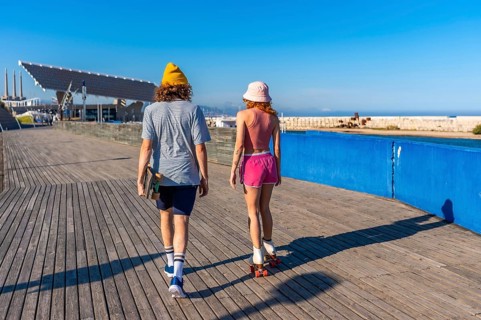 Young man carrying a skateboard strolls alongside a roller skating woman on a wooden pier, enjoying