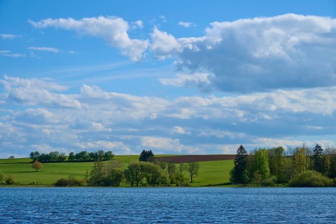 Ober- Moosersee surrounded by green fields and trees under a partly cloudy blue sky, spring,