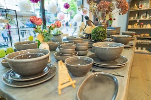 Tableware set in shades of grey on a table surrounded by floral decorations in front of a window,