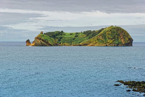 Green, rocky island and protected area Ilhéu de Vila Franca do Campoin a calm sea under a cloudy