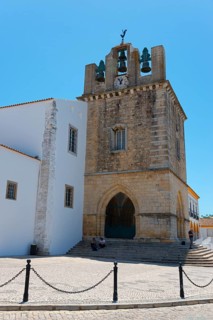 Historic church with bell tower, surrounded by white buildings, under a bright blue sky, Sé
