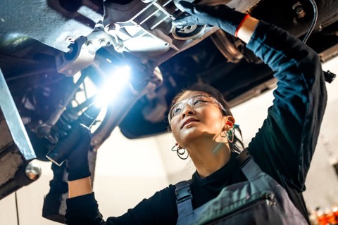 Young woman mechanic wearing safety glasses using flashlight while inspecting car chassis in a