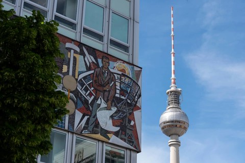 Television tower and building with wall mosaic and tree in front of blue sky, combination of art