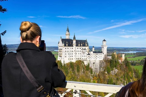 Woman photographing Neuschwanstein Castle in front of a picturesque landscape under a clear sky,