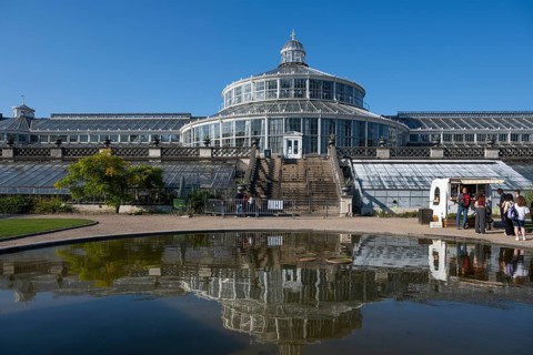 Large historic greenhouse reflected in a pond, palm house with glass facade, blue sky, Botanical