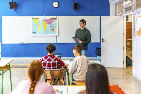 Geography teacher explaining a lesson holding a clipboard and smiling to elementary school students