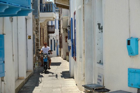 A narrow white alley with blue shutters. A man rides a motorbike through the sun, Mandraki,