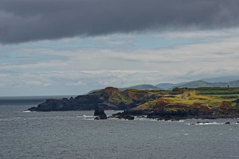 A rocky coastline with green hills in the background under a cloudy sky, Fenais da Luz, Sao Miguel