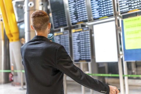 Businessman holding passport and pulling suitcase while checking flight schedule on departure board