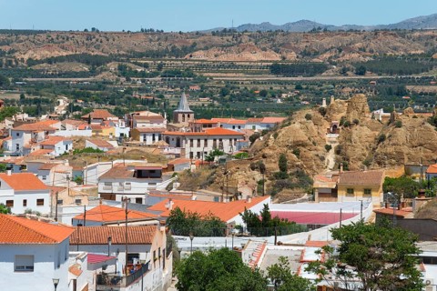 Panoramic view of a village with red roofs and mountains in the background under a clear sky, cave