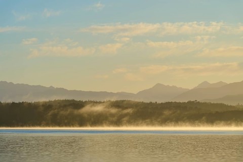 Fog over a calm lake under a clear sky with mountains in the background, Lake Mahinapua, Ruatapu,