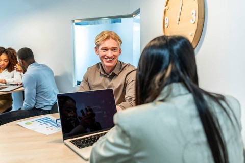 Two managers are smiling and talking during a meeting in a coworking office, using a laptop and