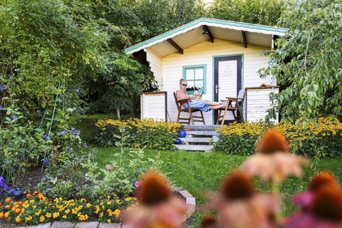 A woman sits relaxed on the small veranda of a garden shed and reads. Blooming summer flowers in