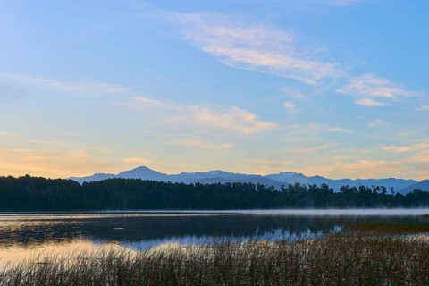 A calm, misty lake in morning sunlight with mountains in the background, Lake Mahinapua, Ruatapu,