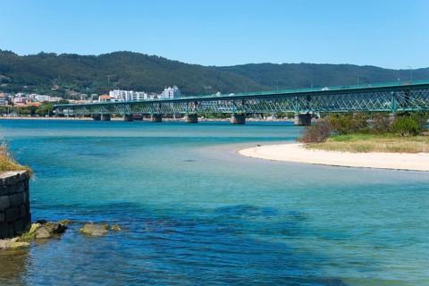 A long bridge crosses a shimmering blue river, surrounded by hills and a town in the background,