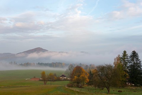 Fog in the valley, surrounded by trees and a small house, with a range of hills in the background,