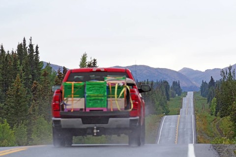 Red lorry with bulky goods drives up and down a straight road lined with forests and mountains,