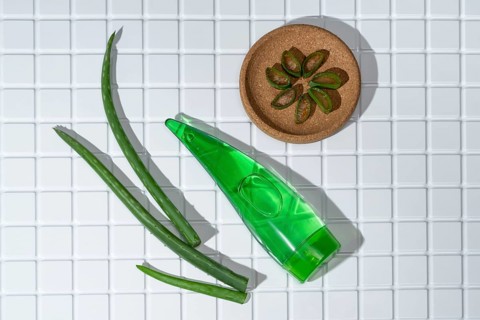 Aloe vera leaves and a container of aloe gel arranged on white bathroom tiles, symbolizing natural