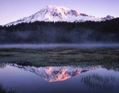 Snow capped mountain reflecting in a calm lake at dusk, surrounded by mist and lush forest. The sun