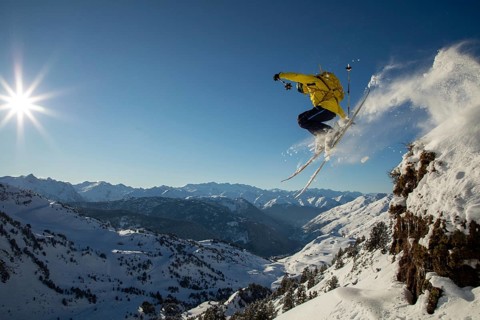 A skier in a yellow jacket performs an exhilarating jump off a snowy cliff, surrounded by a