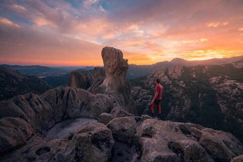 Back view of unrecognizable man standing on rocky outcrop, gazing at vibrant sunset over Pedriza