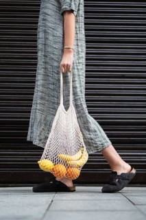 A woman in a long, gray dress carries a reusable mesh bag filled with bananas and oranges. The