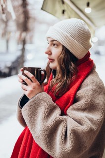 A young thoughtful woman in a white beanie and red scarf holds a warm mug, gazing thoughtfully