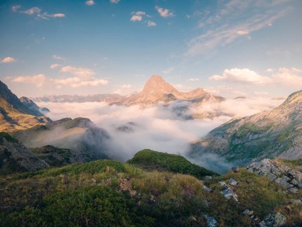 Serene mountain peak rises above a lush green landscape enveloped in wispy clouds under a vast blue