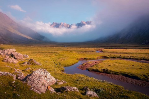 A serene mountain valley landscape with a winding river and mist shrouded peaks in summer. The warm