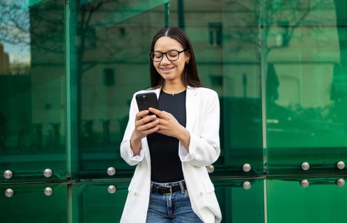 Hispanic businesswoman wearing glasses and a white blazer, smiling while using her smartphone