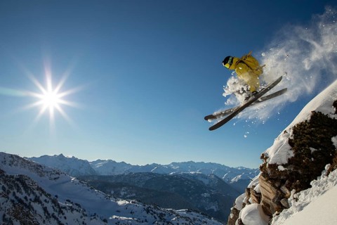 A skier in yellow gear performs a thrilling jump off a cliff in a sunny, snow-covered alpine