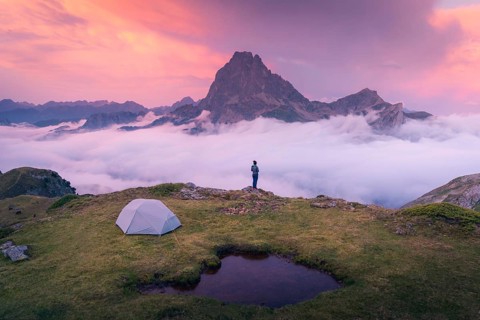 A camper stands near a tent overlooking a majestic mountain peak above a sea of clouds during a