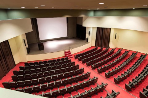 An empty convention center featuring rows of seats facing a wooden stage and a large white screen,