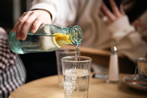 Cropped unrecognizable girl pours water from a bottle with a slice of lemon into a glass in a cafe,