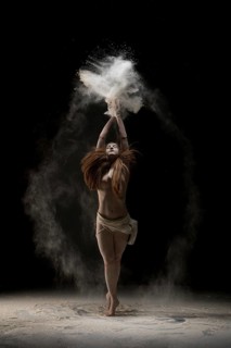 Slim young barefoot woman throwing sand over head while dancing against black background