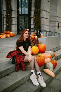 A young woman in autumnal attire sits on stone steps adorned with pumpkins in an urban Montreal
