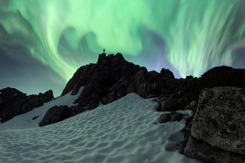 A breathtaking Aurora Borealis illuminates the night sky above a snowy, rugged mountain terrain. An