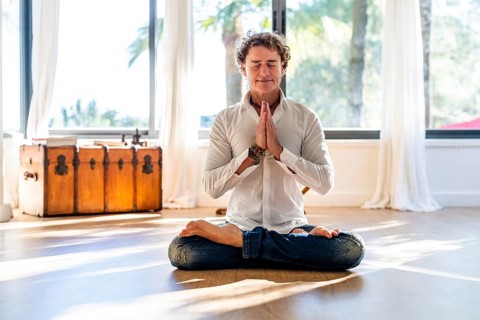 A calm man with curly hair meditates with eyes closed, seated in a lotus pose, hands in prayer