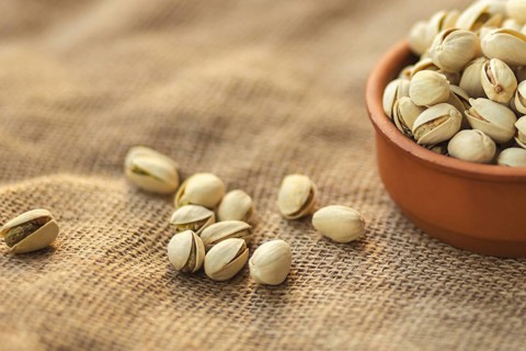 Close up of a clay bowl filled with pistachios on a burlap surface. Several pistachios are