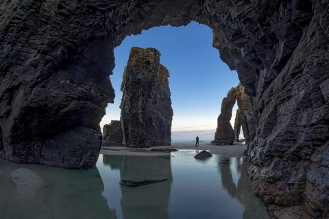 A person stands between towering sea arches, reflected in calm tidal pools, under a serene sky. The