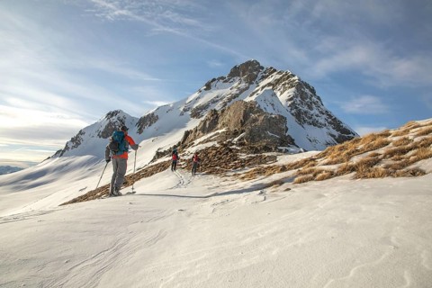 A group of ski tourers makes their way up a snow-covered mountain slope under a clear blue sky. The