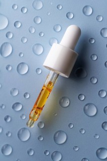 A close-up of a pipette filled with vitamin C serum on a blue surface decorated with water droplets