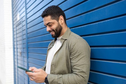 A man stands against a blue wall, smiling as he looks at his smartphone in the city. He wears a